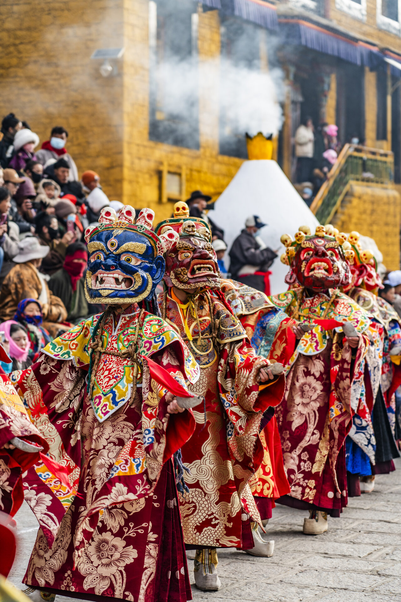 A line of wrathful Guru DrakMar dancers in red costumes process across the courtyard at Tsurphu Monastery.